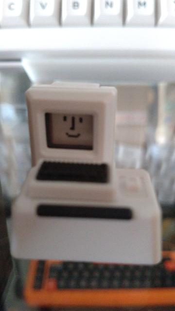 Ein winziger Monitor mit einem Mac-Smiley auf dem "Bildschirm", davor eine winzige Tastatur auf einem richtig fetten Sockel (der eigentlich eine Tab-Taste und im Bild etwas unscharf geraten ist), alles in Computer-Grau-Beige, die Tasten der Tastatur in Schwarz.

Im Hintergrund und durch den Glasboden hindurch sieht man Tastaturen in Orange-Schwarz und Weiß-Schwarz.