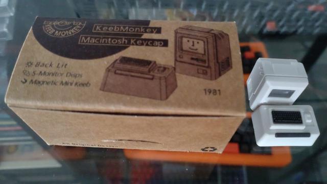 Eine ca. 7cm × 4cm große Schachtel in kartonbraun mit Beschriftung "KeebMonkey Macintosh Keycap" und "1981" sowie einer stilisierten Abbildung ebendieser.

Daneben von oben gesehen ebendiese Keycap bzw. Keycaps:

Ein winziger Monitor mit einem Mac-Smiley auf dem "Bildschirm", davor eine winzige Tastatur auf einem richtig fetten Sockel (der eigentlich eine Tab-Taste und im Bild etwas unscharf geraten ist), alles in Computer-Grau-Beige, die Tasten der Tastatur in Schwarz.

Im Hintergrund und durch den Glasboden hindurch sieht man Tastaturen in Orange-Schwarz und Weiß-Schwarz.