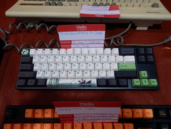 Bild von einem Ausstellungstisch an einem Mechanical Keyboard Meetup. Man sieht drei Tastaturen:

Oben von hinten und nur teilweise sichtbar eine vintage Toshiba "ALPS Bigfoot" Tastatur.

In der Mitte eine Ducky × Varmilo Miya 68 Pro Panda Tastatur mit einer Dwarf Factory The Snoob Bear "Artisan" Tastenkappe in Panda-Farben (weiß-schwarz-grün. Die Beschriftung der alphanumerischen weißen Tasten sieht aus wie Stücke aus grünen Bambus-Stangen. Auf der Leertaste ist eine Szene mit einem Pandabär auf einer Wiese und ein paar Bambussen abgebildet. Die dunkelgrauen Modifier-Tasten haben eine kaum lesbare, schwarze Beschriftung. Enter- und Cursor-Tasten sind hellgrün mit dunkelgrüner Beschriftung. Die Pfeile der Cursortasten sehen aus wie die Spitzen von Bambussprossen.

Unten eine Ducky One TKL mit Orange-Schwarzen Tastenkappen. Man sieht nur die obersten zwei Tastenreihen.

Alle drei Tastaturen haben weiß-rote "Hello, my name is …" Kärtchen zwischen die Tasten geklemmt oder darauf liegend.