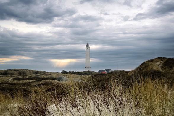 Blåvand's white lighthouse in the distance, surrounded by a cloudy sky and a sun breakthrough in the background, some dunes and marram grass in the foreground.