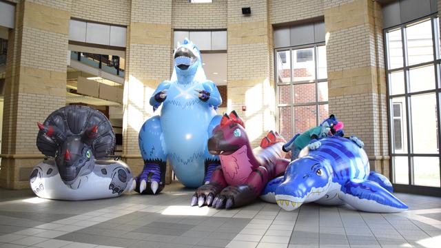 A photograph of an inflatable triceratops tube, standing tyrannosaurus, laying raptor, and pliosaur in a rotunda.  A blue anthropomorphic smilodon in a teal shirt is hugging the pliosaur.