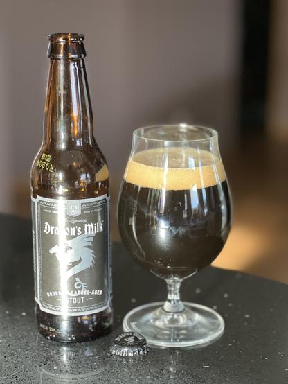 Bright light highlights a bottle of Dragons Milk stout with a black and silver label with a winged dragon in silhouette. A tulip glass of beer is to the right.