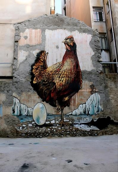 Streetartwall. A beautifully designed mural with a chicken and an egg was sprayed/painted on a still existing old exterior wall of a former house. The brown animal, which is drawn in great detail, stands next to an upright white egg. The sea and icy cliffs can be seen in the background. The surroundings of the backyard buildings and this strange old house wall together with the chicken create a surreal scene. A classic work of street art.