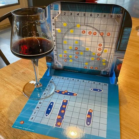 On a table sits a stemmed glass of beer (not much left), beer is amber-brown in color.  Also on the table sits one player's side of the board game 'battleship'.  Table is light oak in color.