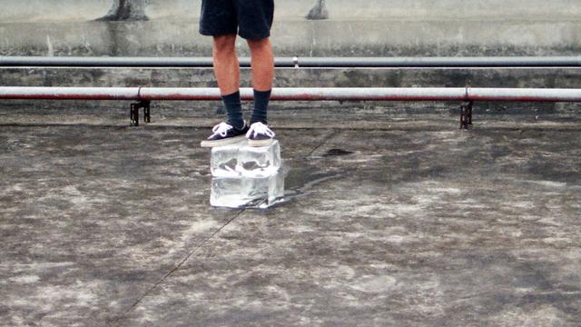 Юный художник неподвижно стоит на кубе из льда и ждёт, когда бруски льда под ним растают и рассыплются. Лица художника не видно. Перформер одет в чёрные просторные шорты, чёрные носки и чёрно-белые кеды. // The young male artist stands motionless on an ice cube and waits for the blocks of ice under him to melt and crumble. The artist’s face is not visible. The performer is dressed in black loose shorts, black socks and black and white sneakers. // Yan-Xiang Lin, “Waiting”, 2016, performance photo documentation