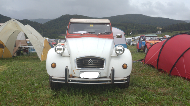 A Citroën 2CV front with white fenders, hood and rolltop roof and a red windscreen frame on an outdoor festival ground with tents and some mountains in the background. The round headlights are on and show bright stripes in the top and bottom third of the headlight glass.