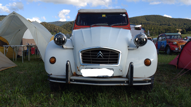 A Citroën 2CV front with white fenders, hood and rolltop roof and a red windscreen frame on an outdoor festival ground with tents and some mountains in the background. The round headlights are dark except for two nearly vertical rectangles close to the left and right rim of the headlights.