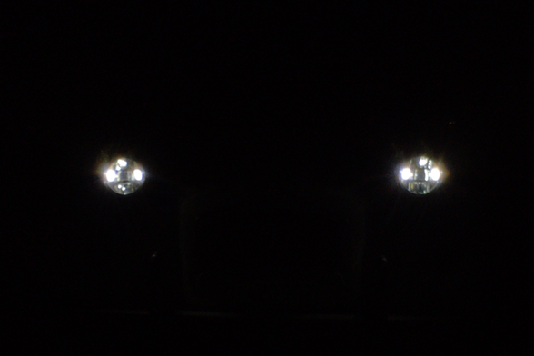 Two LED headlights in the dark. The look like rhombus shaped with round corners and bright spots in all corners but the bottom corner. The kina look like cat eyes.