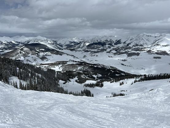 Looking down a steep ski slope, with a valley below and snowy mountains and clouds in the distance.