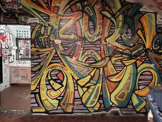 A colorful abstract graffiti style mural to the right of the entrance to one of the restrooms, which is also colorfully decorated with messages and graffiti