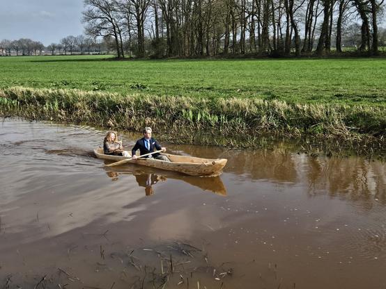 Twee mensen in een kano in het kanaal langs Borger. Achterin de projectleider in Oertijdkleding, halverwege zit ik. We hebben beiden een pedel in de hand.