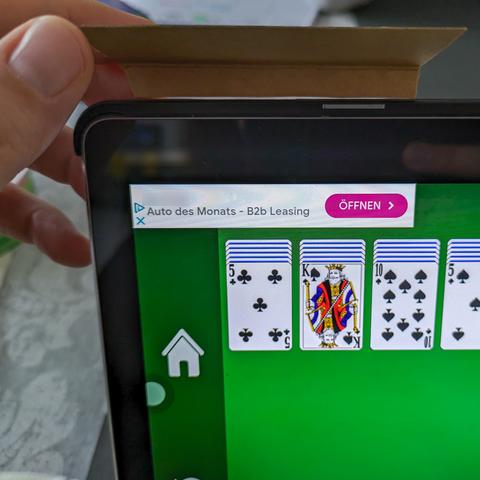 Close-up of the upper left corner of a Huawei tablet. The screen displays a card game with an ad banner in the top left corner. There's a hand holding a piece of cardboard taped to the edge of the tablet.