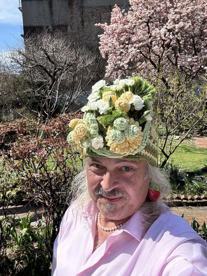 Selfie of someone in a custom crochet hat leaning forward to show the crochet flowers & genuine, in front of a flowering tree in a NYC community garden. Flowering dogwood, side of apartment building, sky, all visible in background.