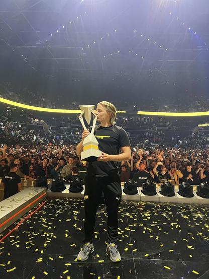 zawodnik Natus Vincere z trofeum Majora na scenie, pozujący do zdjęcia, tyłem do publiczności, całuje trofeum