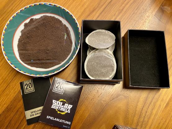 Die Würfel von 20 Strong nehmen unter Klarsichtfolie ein Bad in Kaffeepulver. In der offenen Box liegen einige Kaffeepads. Die Regeln liegen daneben und kommen später noch mit in die Box. Deckel drauf und hoffen…