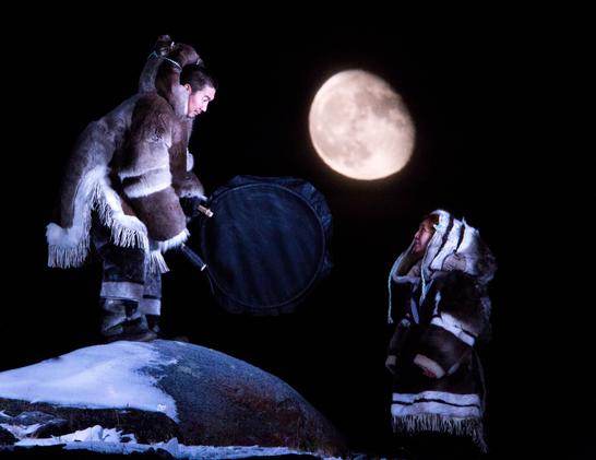 Two Inuuk. A man dressed in caribou clothing drums, standing on a snowy rock. He looks down towards a woman gazing back at him. She is also dressed in skin clothing. Behind them the moon rises in the dark sky.