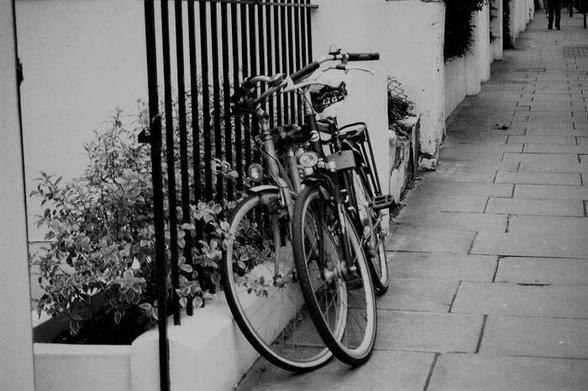 Dos biciletas, apoyadas la una junto a la otra en una verja, en la calle. Imagen en blanco y negro.