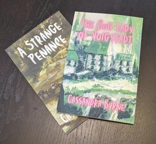 A pair of books displayed on a table; "The Bug Lady of Volgstadt" in pink and green, and "A Strange Penance" in white and brown.