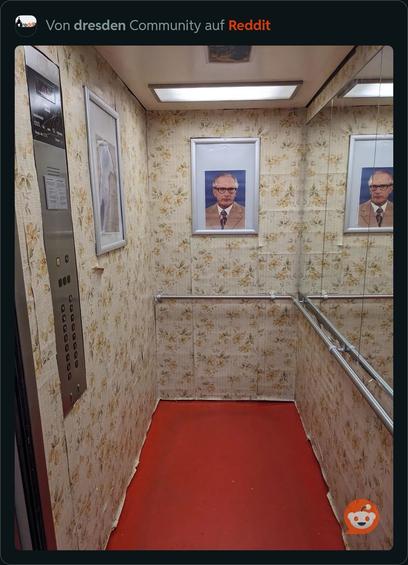 Innenraum eines Aufzugs mit einer alten Blumentapete, einem roten Teppichboden und einem Bild von Erich Honecker an einer Wand. An der linken Wand befindet sich eine Schalttafel mit den Fahrstuhl-Tasten.