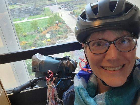 Photo of a smiling white woman with multicolored tortoiseshell glasses wearing a black bike helmet and looking into the Cabrera. Behind her, handlebars of a bike with a blue bag on the front and multicolored streamers hanging from the center of the handlebars.