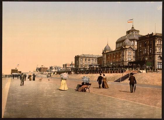 The image appears to be a vintage postcard depicting an early 20th-century scene in Scheveningen, Holland. The architecture and clothing styles suggest it could be from around the turn of the century. The Kursaal is visible on the right side of the postcard, which was likely a popular entertainment venue during this time period. In the foreground, there are pedestrians dressed in period attire, some holding parasols or wearing hats, indicating that the scene captures a leisurely day out in what appears to be a seaside resort. The image is rendered in a soft color palette, with earth tones dominating the composition, which lends a nostalgic atmosphere to the depiction of this bygone era.