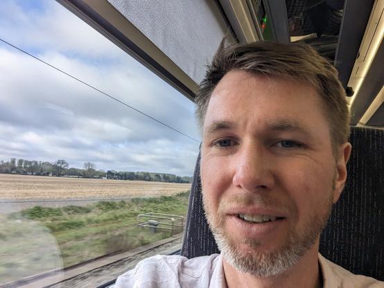 A man with short hair and a stubble beard smiles slightly while taking a selfie inside a train. He wears a light-colored shirt and is seated by the window, through which a rural landscape with open fields and sparse trees can be seen passing by. The sky is partly cloudy.