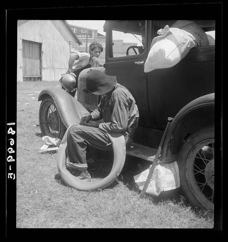 The image is a black and white photograph capturing a scene from an era where tobacco sharecroppers were prevalent in the American South. In the foreground, there's a man sitting on the ground next to an old car. He appears to be resting or perhaps taking a break, as he has one leg crossed over the other and his body leans against the side of the vehicle. The car is an older model, suggesting that this photo was taken in the early 20th century.

The setting seems rural, with dirt ground underfoot and wooden structures visible in the background, which aligns with the sharecropper theme suggested by the textual information provided. There are also other people present in the image, although they are not the primary focus of the scene. Their presence suggests that this might be a communal gathering or rest area for workers.

The overall mood of the photograph is candid and informal, giving us a glimpse into the everyday lives of these individuals, their environment, and their mode of transportation during the time when tobacco farming was a significant part of life in regions like Douglas, Georgia.
