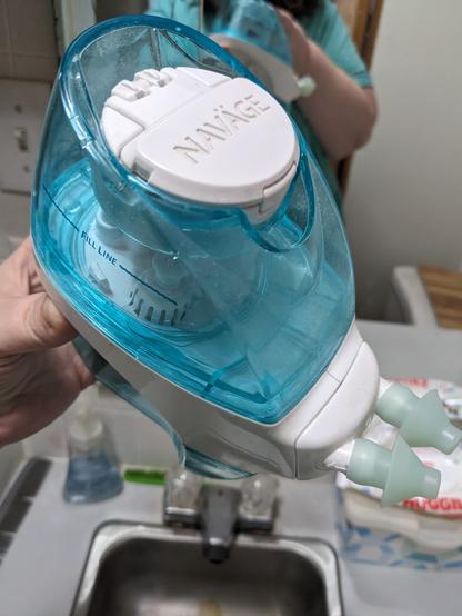 A Navage nasal cleaner. The device has a white and translucent cyan color scheme, with a removable reservoir on the top and bottom of the relatively thin main body. Two silicone ports poke out from the front, intended to go in your nostrils. A bathroom sink and mirror are visible in the background.