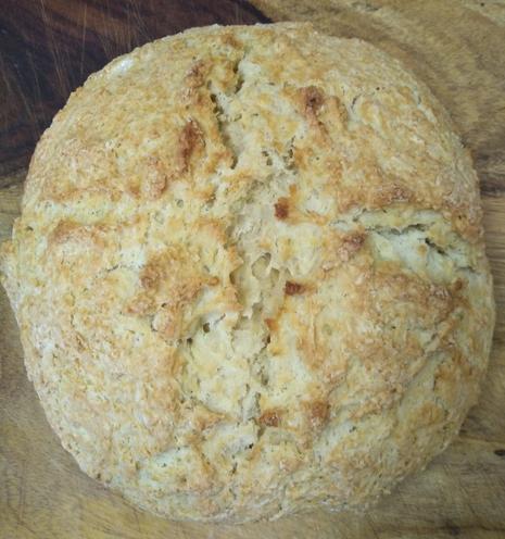 A craggy, golden brown Irish Soda bread round with a cross shaped score across the top sits on a wooden cutting board.
