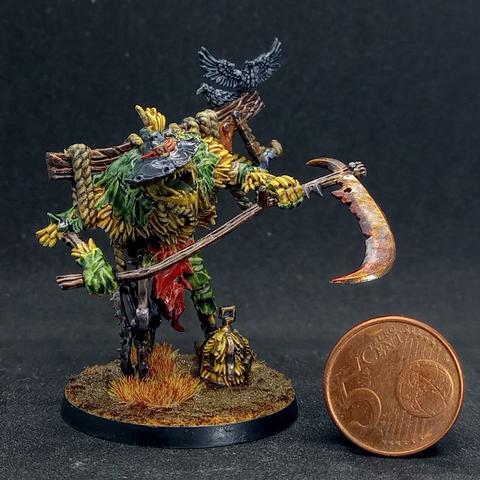 Plastic miniature of an evil animated scarecrow wielding a rusty, slightly bloodied scythe. There are 2 corvids hanging around on its wooden crossbeam.