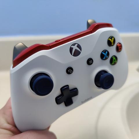 An Xbox One controller made in Xbox Design Lab to color accurately match Sudaksis's color scheme. White controller body with red bumpers and red strip at the top, blue joysticks, metallic blue D-pad, face buttons with black background and colored letters, and metallic gold triggers. "Sudalsis" is engraved near the bottom.