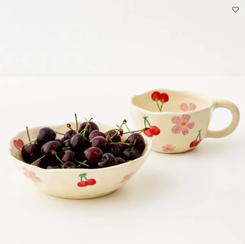 A product photo of a bowl of cherries and an empty tea cup, both vessels are very rustic in form, as if someone has shaped them by hand without caring too much about smoothness but I think they have been slip cast for mass production. Both are cream coloured with "hand painted" cherries and flowers.