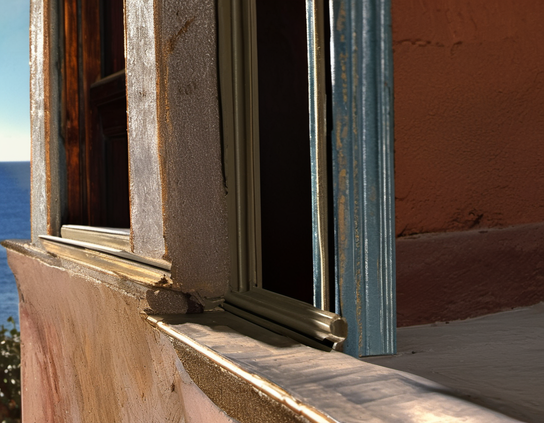 Sunlight glints off metallic paints on a textured wall with open windows by the sea, in the process of renovation.