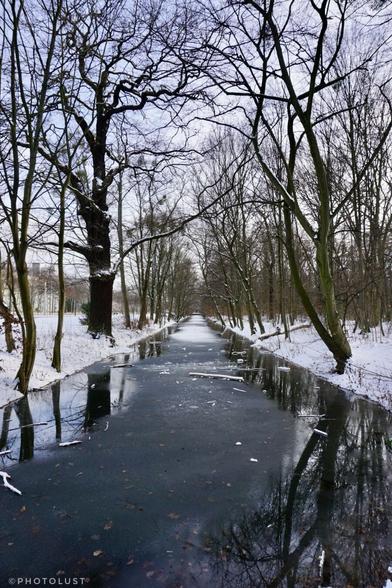 Farbfoto einer Winterszene im Großen Garten zu Dresden. Ein Wassergraben, teilweise mit Eis bedeckt. An den Ufern kahle Bäume. Der Boden ist schneebedeckt.