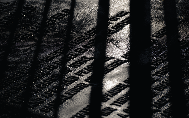 This is a black and white photograph.

Railings that are out of shot are casting hard shadows across a memorial stone laid on the ground.