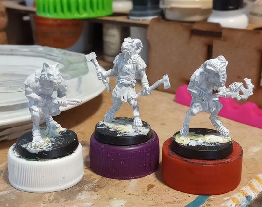 3 metal miniatures of animal people attached to bottlecaps, primed white. The two goat people have axes and the wolf person has a shotgun.