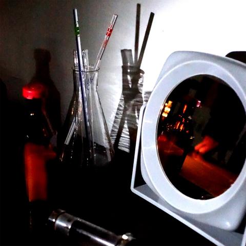 Auf einem Schreibtisch stehen im Halbdunkel: Ein weißer Plastikspiegel, in dem sich die anderen Gegenstände und eine Hand spiegeln, ein Medikamentenfläschchen mit weißem Inhalt und ein Glasbehälter mit mehreren Pipetten.