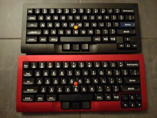 Two Tex Shura 60% keyboards with a 0.89u keycaps cursor cluster in the bottom right corner. They both have sculped, vintage looking white on black keycaps with legends in a typewriter font. They have a thumbstick between the G, H and B keys like ThinkPads and the black keys as mouse buttons in front of the spacebar, embedded in the at that position lowered frame.

Differences:

Top one has an officially black but actually more anthracite metal frame, bottom one a cold red frame.

Top one has an orange thumbstick, bottom one a red one.

Top one has an IBM-blue Enter key, bottom one a black one.

Top one's cursor cluster has just the four cursor keys in an upside down T layout, bottom one has two additional blank keys and hence a 3x2 layout.