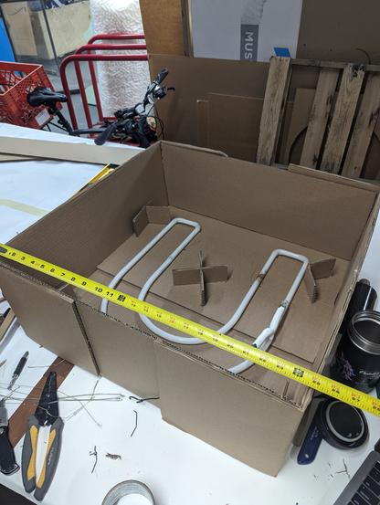 A large neon letter "U" wire tied into a layer of cardboard with stacking cardboard supports for layers in another cardboard box. A tape measure in front reads 25 inches along the square box.