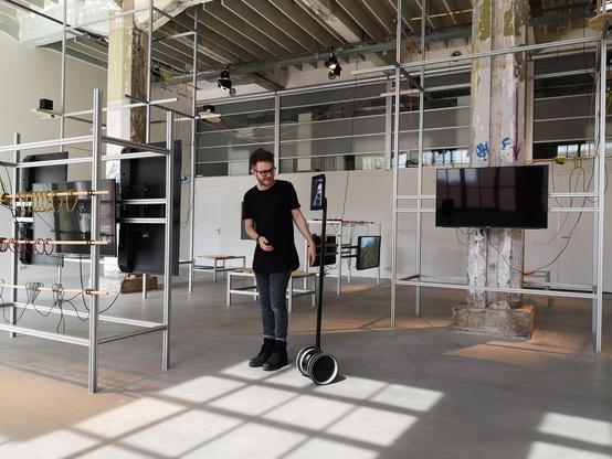 A big exhibition room occupied by bare metal structures that hold TVs in all directions. A person in the center is looking at a tablet screen attached to a pole with wheels. The tablet has the face of a person looking at these tvs.