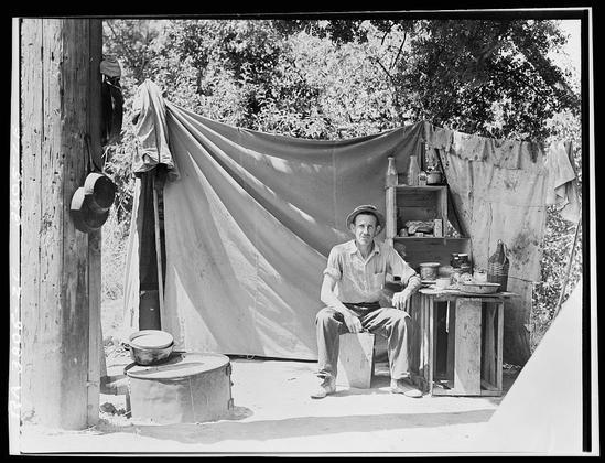 The image appears to be a black and white photograph featuring an individual sitting outdoors. The person is seated on what looks like a wooden bench or table, with their legs crossed and hands resting on their knees. They are wearing a light-colored shirt and dark pants, which suggests casual attire suitable for the outdoors.

In the background, there's an old-fashioned campfire setup with what appears to be a tarp or sheet providing some shelter from the elements. There are also other items scattered around, including bottles, bowls, and a hat hanging on a wooden post. The setting is rustic and natural, indicative of a rural or wilderness environment.

The photograph has a vintage quality to it, suggesting that it might be from an earlier era in photography, possibly mid-20th century given the style of clothing and equipment present. There's no visible text on the image to provide additional context or information about the person or location.