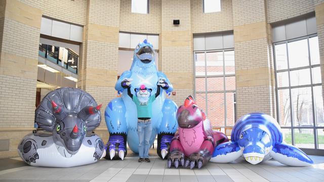Four oversized inflatable dinosaurs in a brightly lit atrium: a grey triceratops tube, a blue feathered tyrannosaur, a maroon feathered utahraptor, and a blue pliosaur.  A blue sabertooth in a teal t-shirt stands in front of the tyrannosaur with his arms raised.