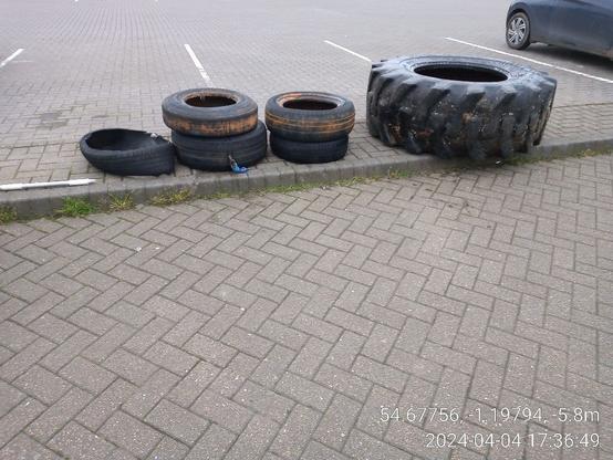 Vor einem Parkplatz liegen teilweise gestapelt 5 Autoreifen und daneben der große Reifen.
