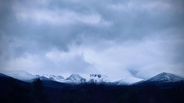 A very monochrome and high contrast view of the Cairngorm mountains taken at dusk. The foreground is almost black and features silhouetted trees, behind that are several snow-covered mountains. The sky is very moody with dark clouds.