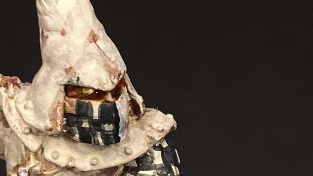 Close-up of a figurine with a white hood and a half black and white chequered mask.