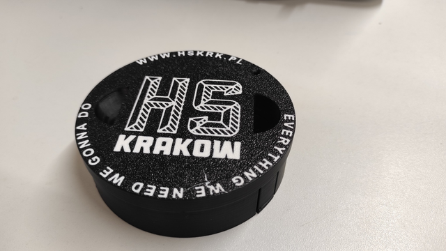Exterior of the projector lens cap, the front-facing wall features the printed logo of Hackerspace Kraków, surrounded by the text - at the top 'www.hskrk.pl' and at the bottom 'everything we need we gonna do.' The print was most likely done on the textured sheet of the 3D printer table, which can be inferred from the specific roughness.