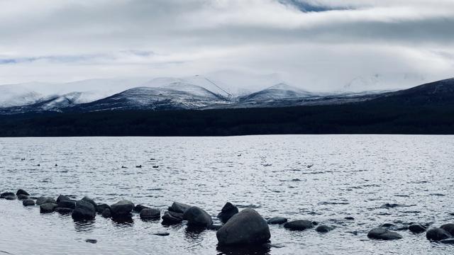 A mostly monochrome photo of Cairngorm on the far side of Loch Morlich. The bottom half of the picture is the loch, there is a line of large boulders in the water in the foreground. Beyond the loch are some dark mountains, the tops have a good dusting of snow. The middle mountain is Cairngorm and the (currently closed) funicular railway can be seen if you zoom in.