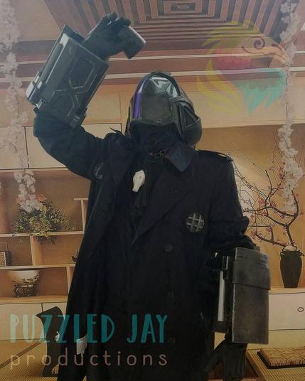Four pictures of a man cosplaying as Bondrewd from Made in Abyss. The character is dressed in a black overcoat with dark metallic gauntlets, greaves, and helmet. The helmet has a glowing, purple slit running vertically down the middle of the helmet.