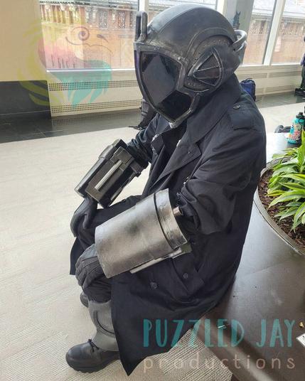 Four pictures of a man cosplaying as Bondrewd from Made in Abyss. The character is dressed in a black overcoat with dark metallic gauntlets, greaves, and helmet. The helmet has a glowing, purple slit running vertically down the middle of the helmet.