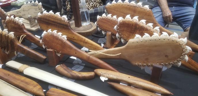 A table filled with hand made Hawaiian clubs. They are long and flat like a paddle, they are made from wood and sharks teeth all around the edges.